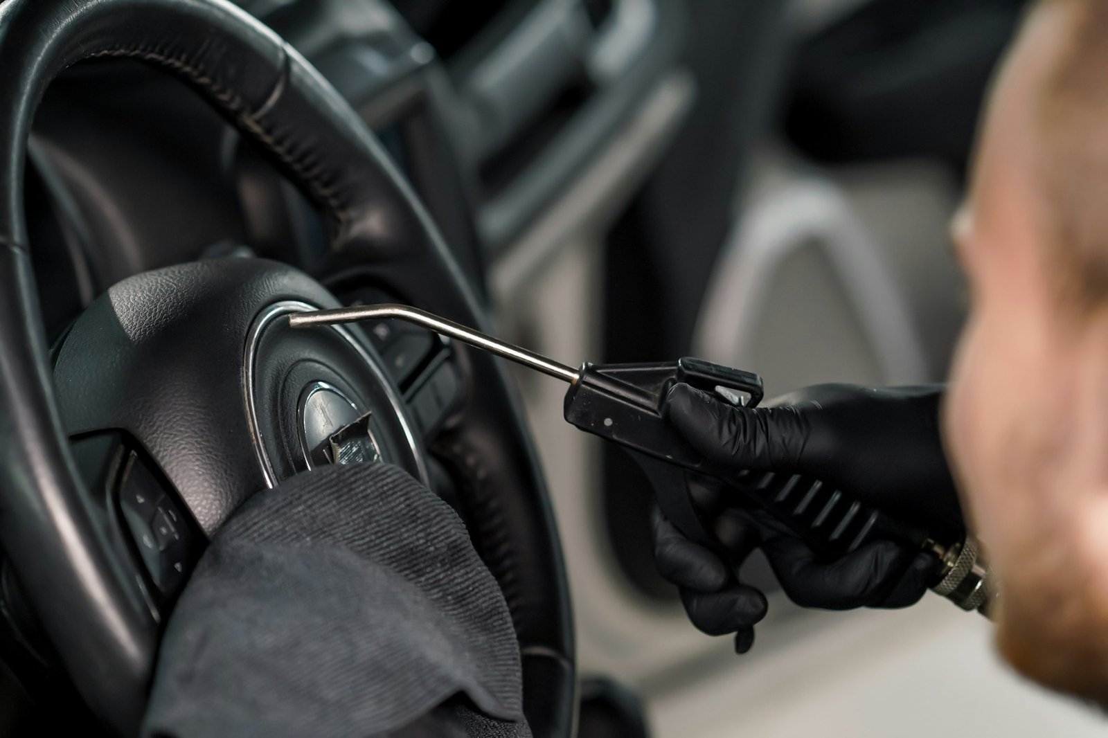 A worker using a blow gun and rag to clean a car's steering wheel during detailing.