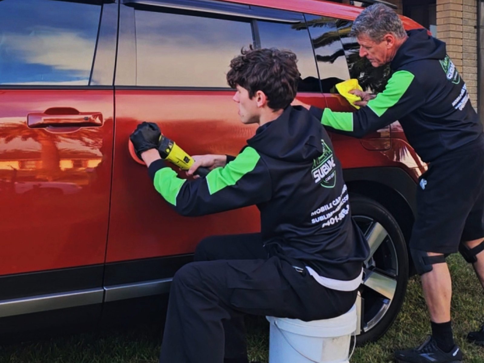 Two detailers working on polishing the side of a red SUV during a detailing service.