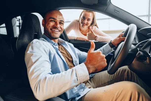 Smiling man in a car giving a thumbs-up with a woman giving a thumbs-up outside the window.